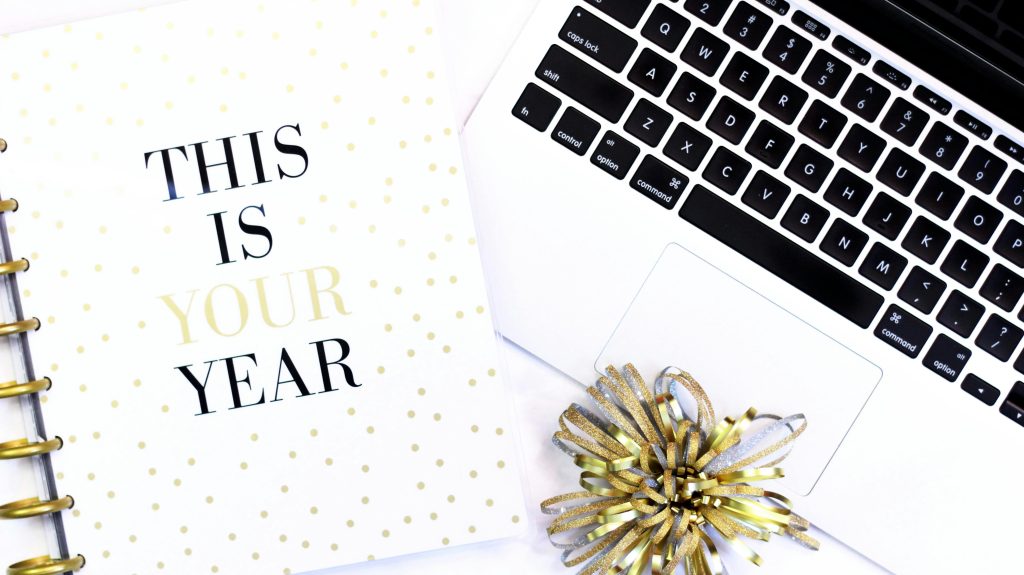 Spiral notebook that says "This is your year" in all capital letters laying next to an open MacBook .