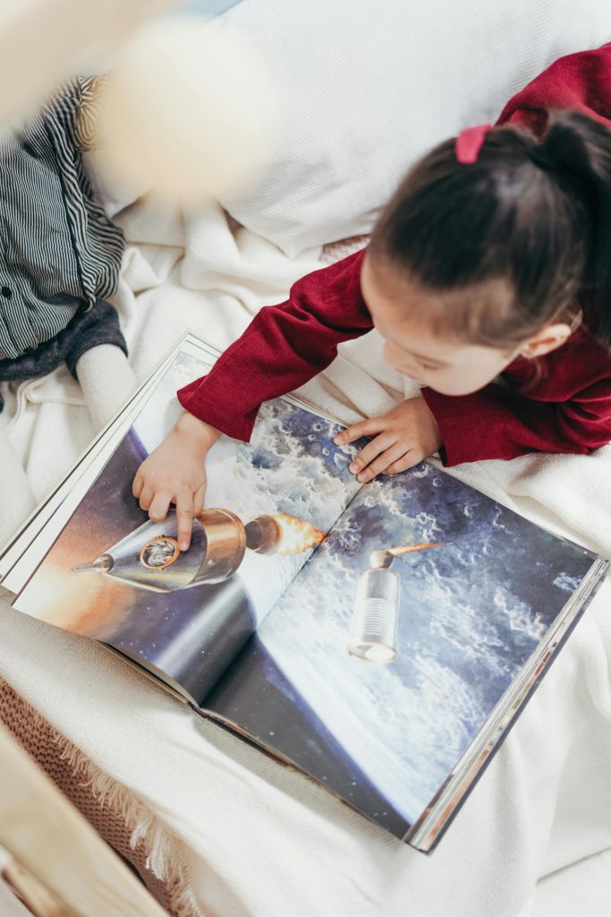 Image of a young girl looking at a book about space and pointing to an image of a satellite.