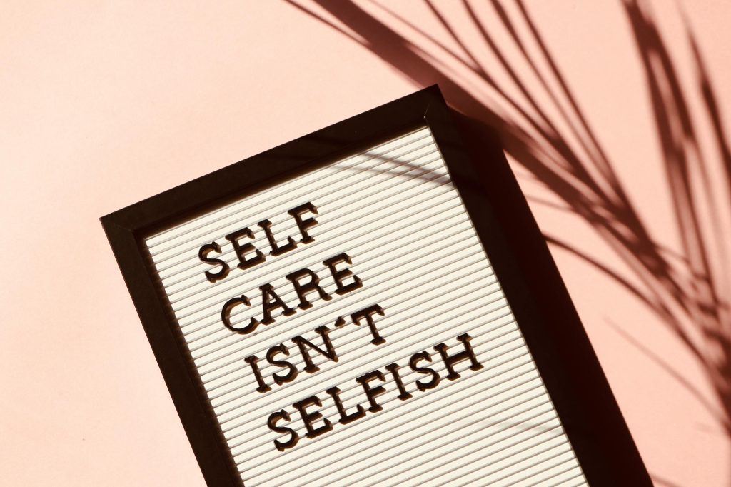 White board with black frame and black letters that says "self care isn't selfish" in front of a pink background.